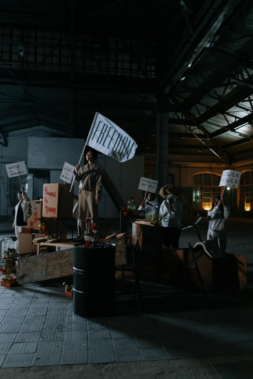 a group of people holding signs in a warehouse, inspired by Vanessa Beecroft, unsplash, conceptual art, unreal engine. retro film still, soviet yard, [ theatrical ], freedom