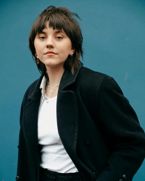 a woman standing in front of a blue wall, an album cover, inspired by Ion Andreescu, trending on pexels, wearing black overcoat, wearing collar on neck, promotional image, rebecca sugar