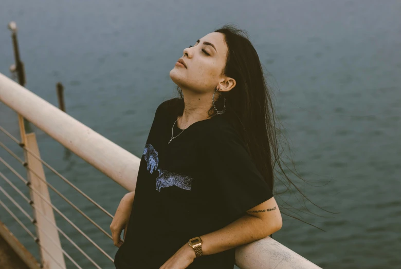 a woman standing on top of a bridge next to a body of water, inspired by Elsa Bleda, trending on pexels, aestheticism, wearing black tshirt, wearing a native american choker, windblown dark hair, sitting