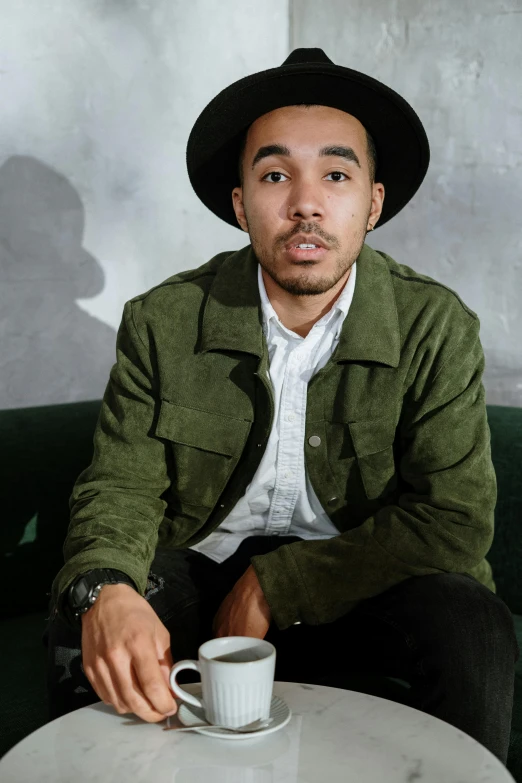 a man sitting on a couch with a cup of coffee, by Stokely Webster, wearing a hat, ashteroth, portrait pose, liam