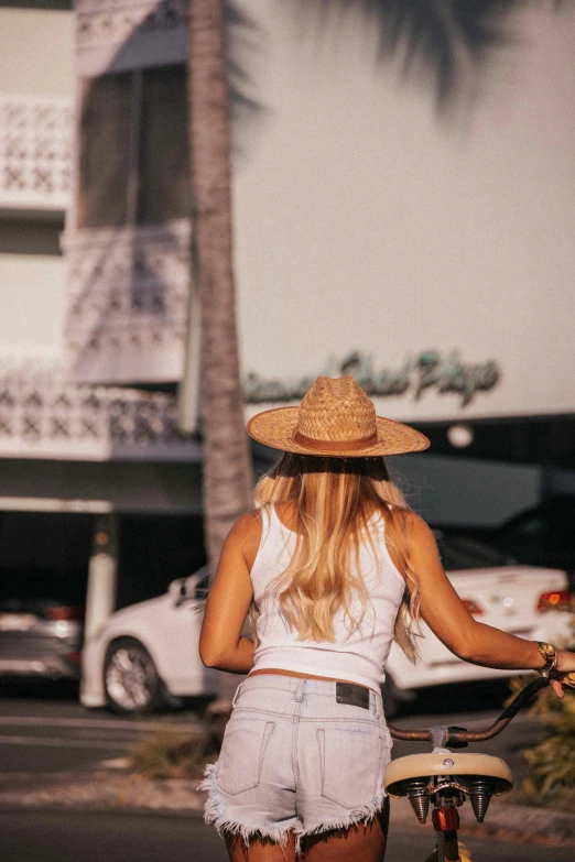 a woman riding a bike down a street next to a palm tree, white straw flat brimmed hat, wearing a crop top, back view also, standing in a parking lot