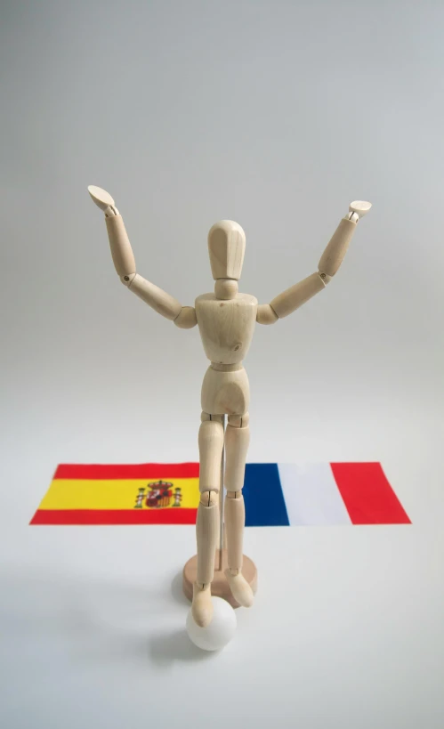 a wooden mannequin standing in front of a spanish flag, arms stretched out, five points of articulation, french flag, two legs two arms one head