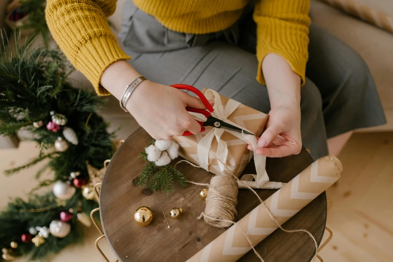 a woman sitting on top of a wooden table holding a pair of scissors, by Julia Pishtar, pexels contest winner, birthday wrapped presents, weaving, christmas, golden ribbon