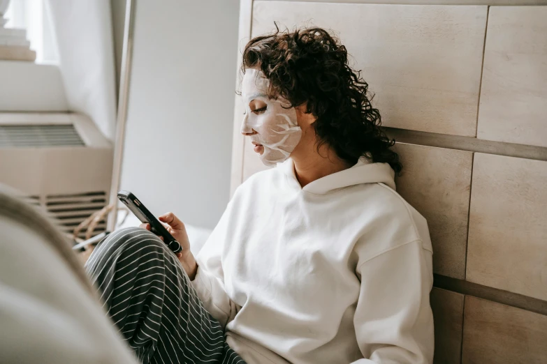 a woman sitting on a bed using a cell phone, trending on pexels, face mask, white facepaint, bedhead, thick jawline