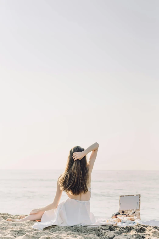 a woman sitting on top of a sandy beach next to the ocean, unsplash, conceptual art, brown colored long hair, on a white table, arms behind back, sunfaded