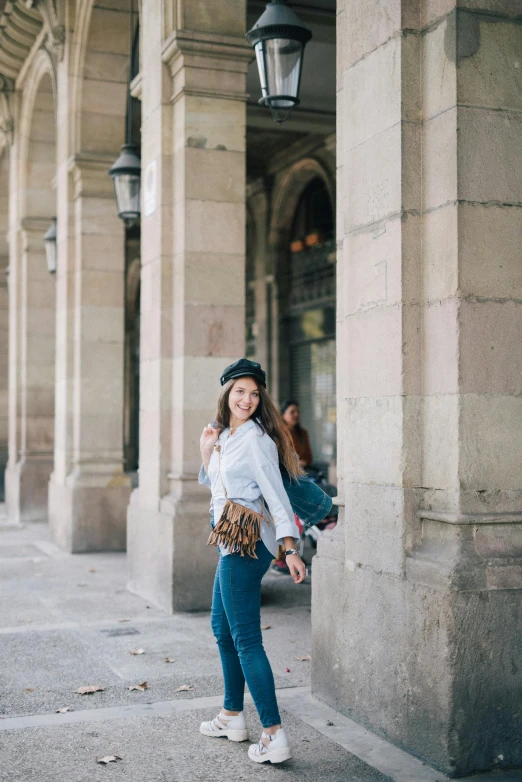 a woman standing in front of a building, inspired by Luis Paret y Alcazar, trending on pexels, wearing jeans, girl with brown hair, with hat, smiling