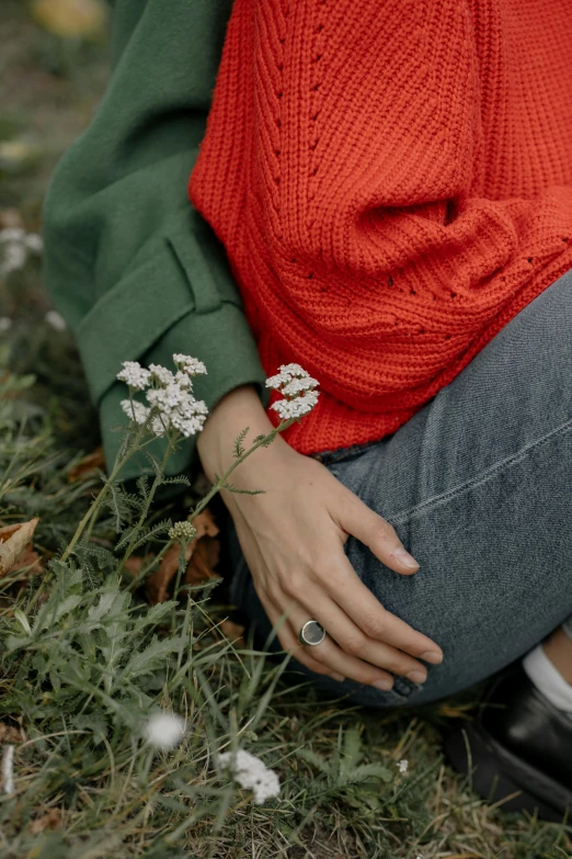 a woman sitting on top of a grass covered field, inspired by Elsa Bleda, trending on pexels, red sweater and gray pants, rings, with flowers and plants, foliage clothing