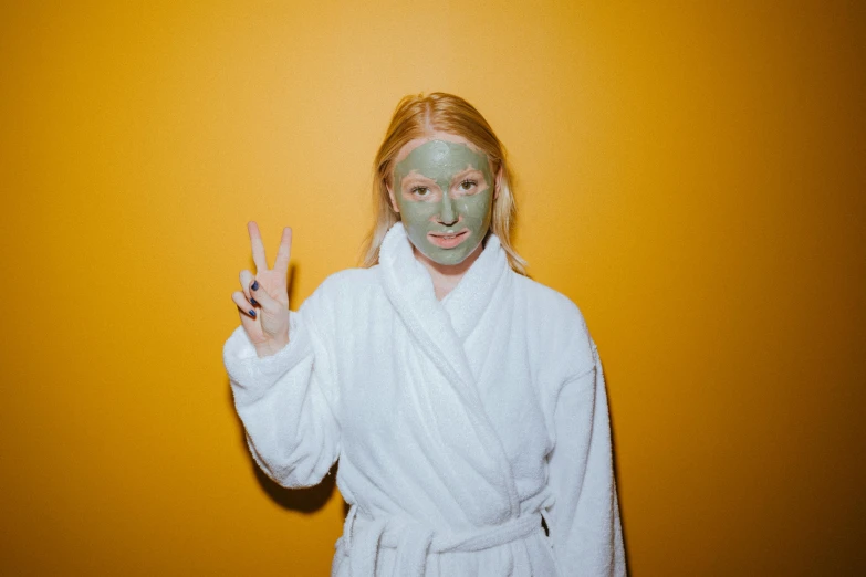a woman in a bathrobe making a peace sign with her hands, inspired by Elsa Bleda, trending on pexels, green facemask, mud on face, yellow skin, a blond