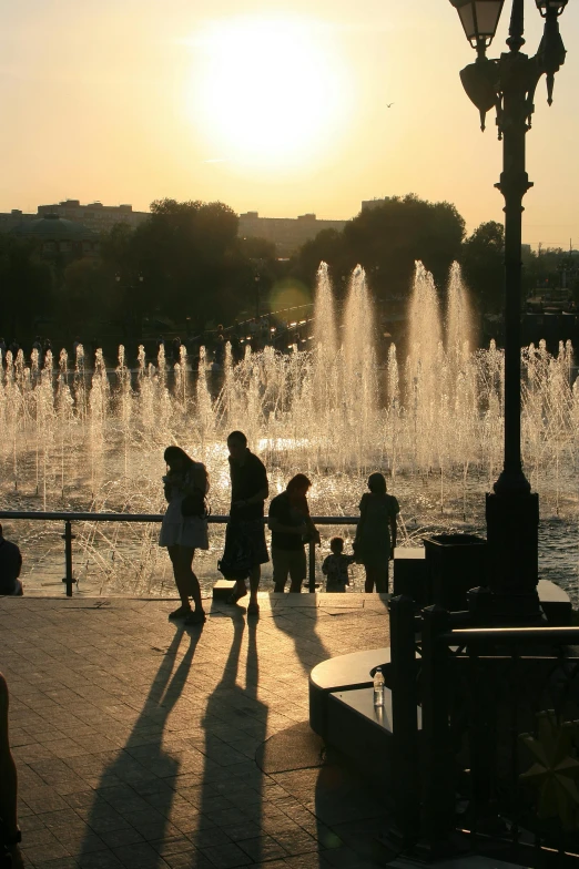 a group of people standing in front of a fountain, during a sunset, moscow, full morning sun, bustling