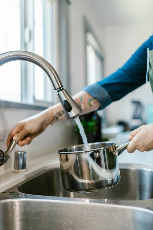 a woman pouring water into a stainless steel sink, by Everett Warner, pexels, tattooed, pots and pans, caucasian, home setting