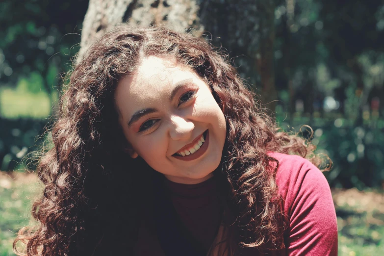 a beautiful young woman sitting in front of a tree, a portrait, by Gwen Barnard, pexels contest winner, dark short curly hair smiling, portrait sophie mudd, official screenshot, curly red hair