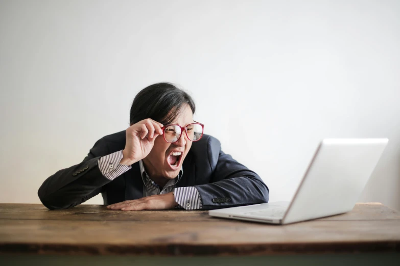 a man sitting at a table in front of a laptop, by Julia Pishtar, pexels, people screaming, wearing small round glasses, asian male, horrified