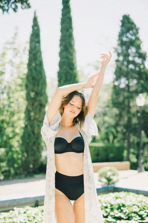 a woman in a bikini and cardigan posing in front of a fountain, a picture, by Julia Pishtar, unsplash, arabesque, posing together in bra, with arms up, wearing black silk robe, in garden