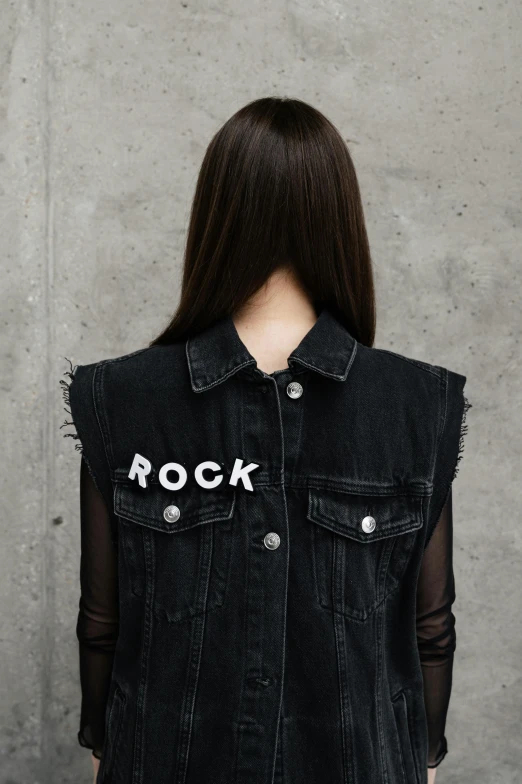 a woman standing in front of a concrete wall, an album cover, by Robbie Trevino, black vest, sticker of a rock band, detail shot, denim