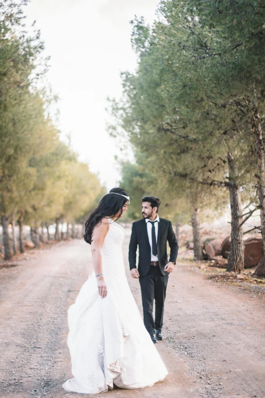 a bride and groom walking down a dirt road, arab ameera al-taweel, trees in the background, wine, in front of white back drop