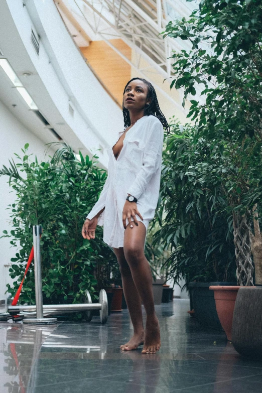 a woman in a bathrobe standing next to a potted plant, by Matija Jama, unsplash, happening, wearing a wet white short dress, dark skinned, in a mall, badass pose