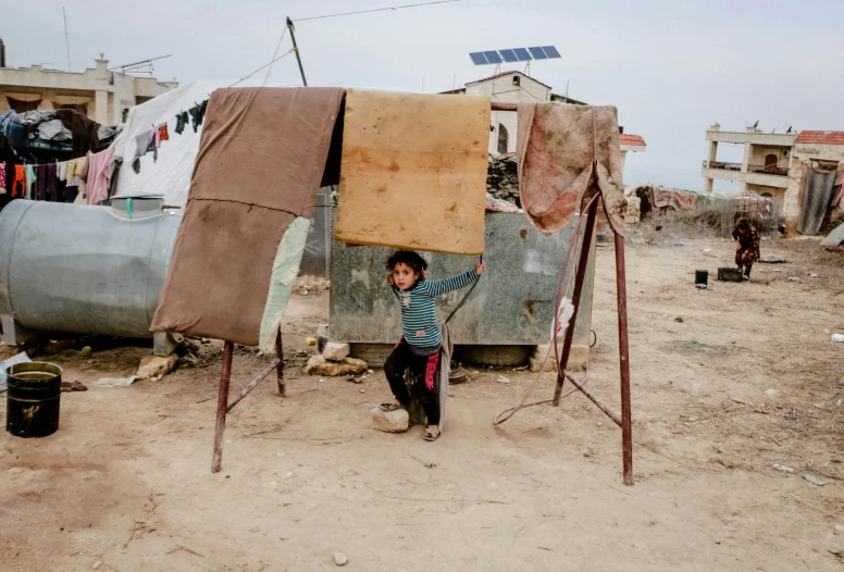 a little boy that is standing in the dirt, by Nathalie Rattner, makeshift houses, holding up a large shield, shutter, amr elshamy