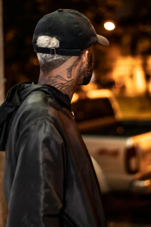 a man standing on the side of a street at night, a tattoo, trending on pexels, photorealism, wearing a backwards baseball cap, slicked-back hair, profile image, black hood