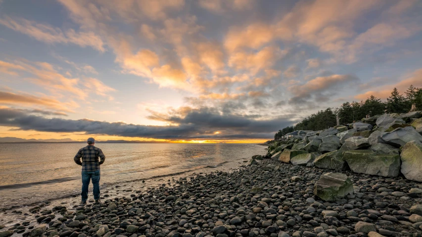 a man standing on a rocky beach at sunset, by Terese Nielsen, pexels contest winner, british columbia, sunset + hdri, sunset and big clouds behind her, fishing