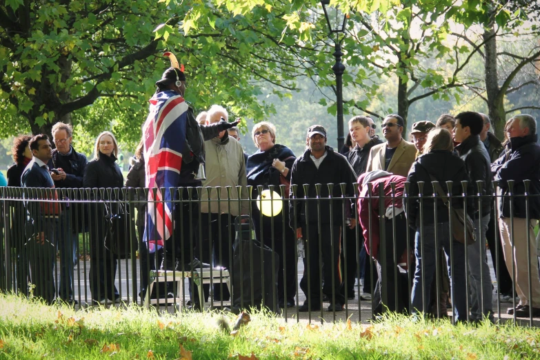 a group of people standing next to a fence, flickr, private press, british flag, a park, giving a speech, square