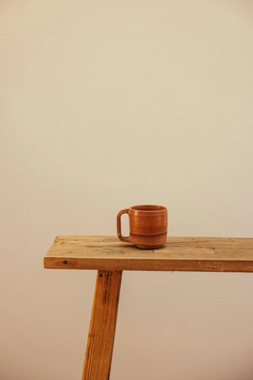 a coffee cup sitting on top of a wooden table, by David Simpson, mingei, terracotta, minimalistic, medium long shot, bench