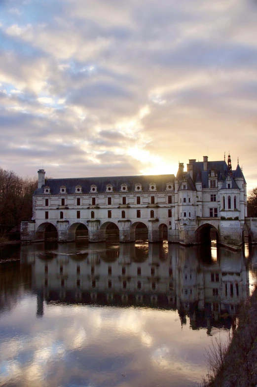 a castle sitting on top of a river next to a bridge, an album cover, inspired by Jean Tabaud, flickr, baroque, sun down, château de cheverny, seen from outside, les catacombes