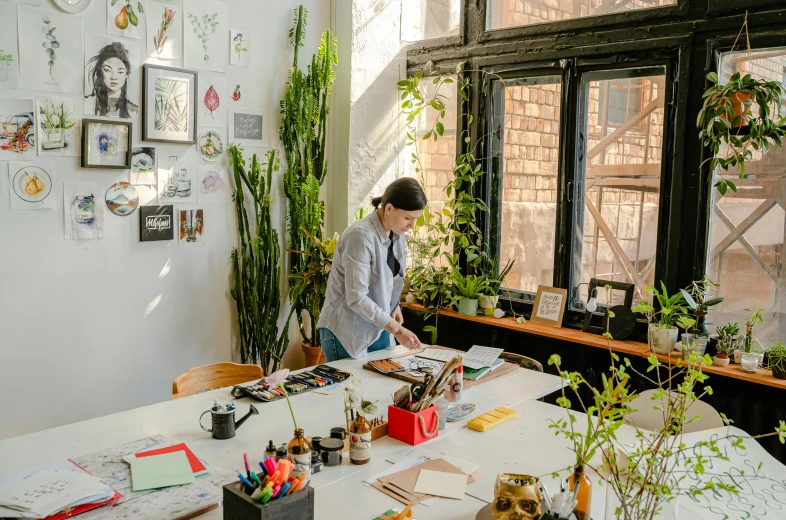 a woman sitting at a table in front of a window, pexels contest winner, arbeitsrat für kunst, room full of plants, drawn projects on paper, set inside of office, crafts and souvenirs