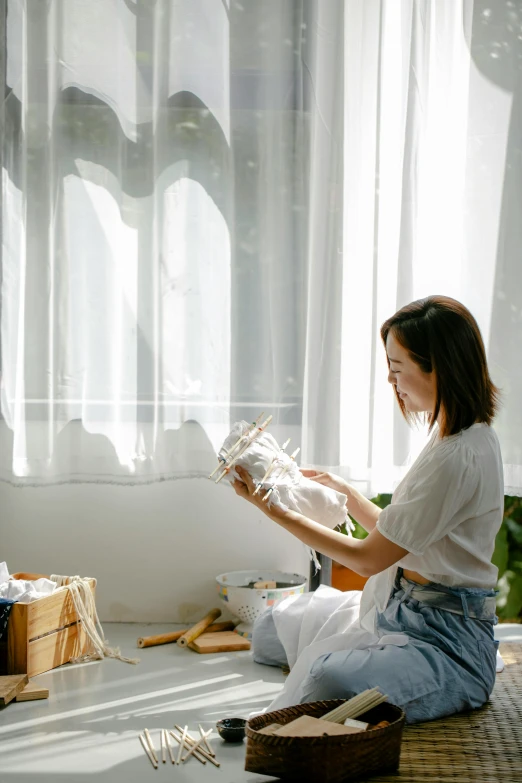 a woman sitting on the floor in front of a window, japanese collection product, white apron, clear painting and good lighting, weaving