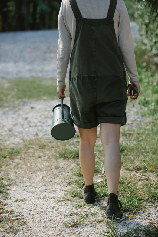 a woman walking down a dirt road holding a watering can, overalls, forest green, short, subtle details