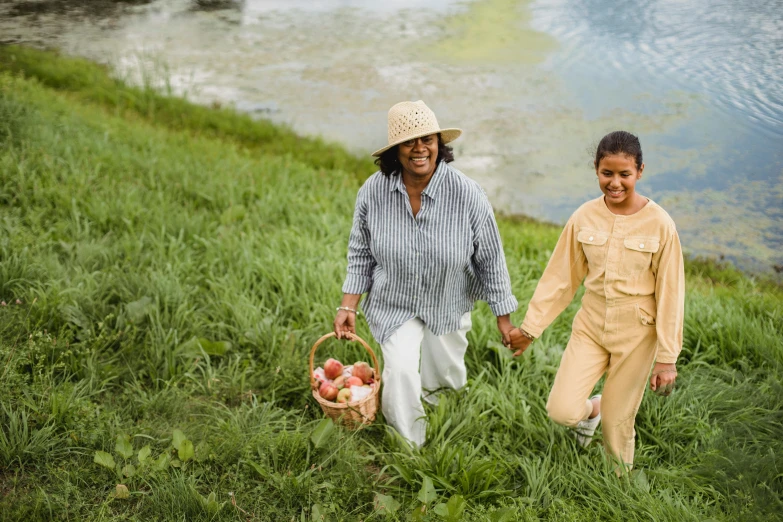 a couple of people that are walking in the grass, a fruit basket, wearing farm clothes, at the waterside, family friendly