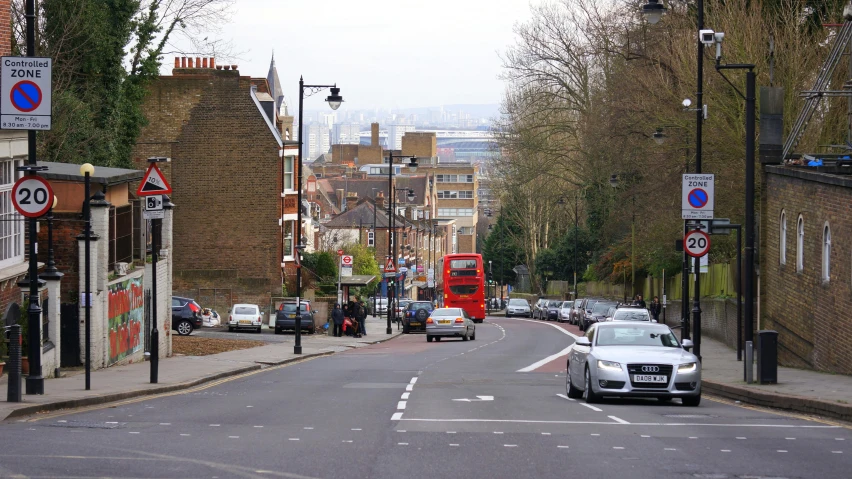 a red double decker bus driving down a street, a photo, highgate cemetery, view from far away, sloped street, 2000s photo