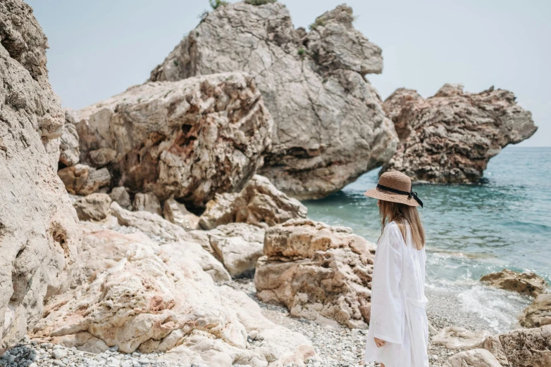 a woman standing on a rocky beach next to the ocean, by Emma Andijewska, pexels contest winner, wearing a white hospital gown, cyprus, wearing a travel hat, florence pugh