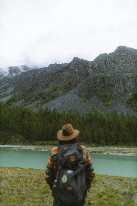 a man with a backpack standing in front of a lake, by Anna Haifisch, wearing a travel hat, in a mountain valley, coloured photo, low quality footage