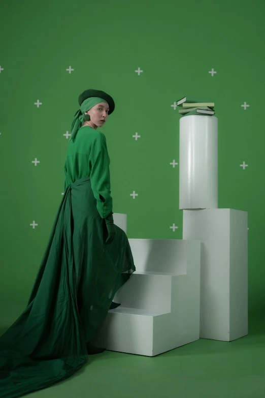 a woman in a green dress standing next to a stack of books, an album cover, inspired by Art Green, trending on pexels, futuristic fashion show, green hat, green screen, holly herndon origami statue