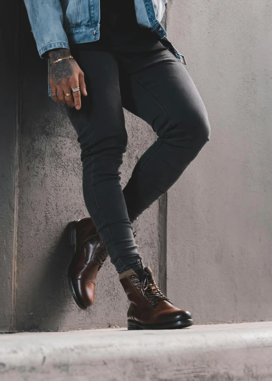 a man in a denim jacket leaning against a wall, a picture, by Carey Morris, trending on pexels, renaissance, wears brown boots, detailed product image, background image, black leather shiny jeans