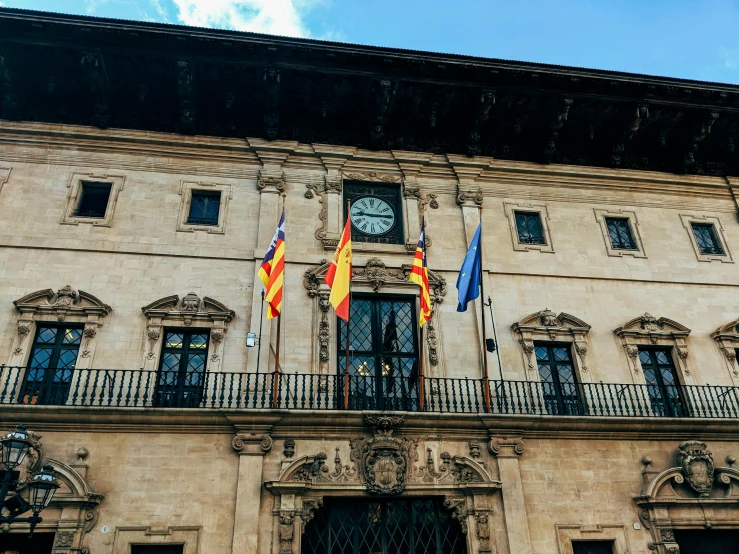 a clock that is on the side of a building, a cartoon, by Tomàs Barceló, pexels contest winner, art nouveau, flags, in balcony of palace, albert ramon puig, thumbnail
