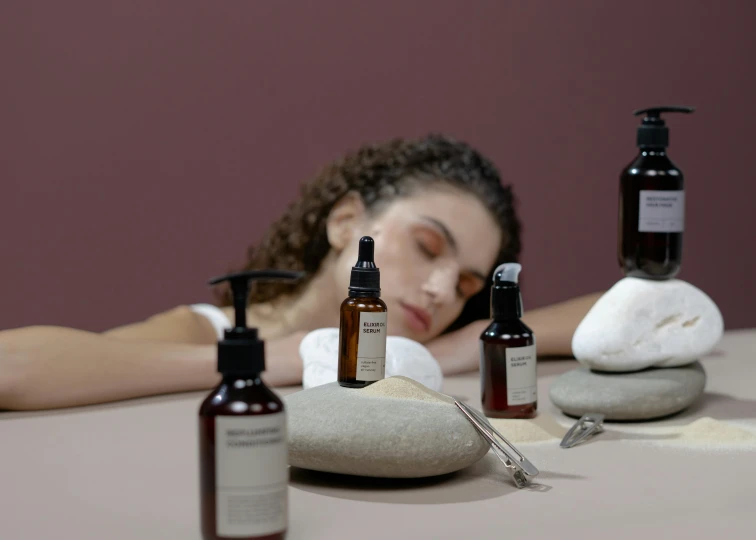 a woman laying on top of a table next to bottles of skin care, a still life, by Emma Andijewska, trending on pexels, mannerism, ultrarealistic, brown, bedhead, middle eastern skin