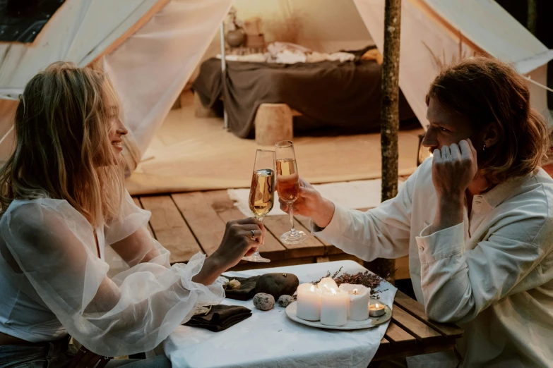 a couple of women sitting at a table with a glass of wine, pexels contest winner, romanticism, glamping, couple on bed, champagne commercial, eating outside