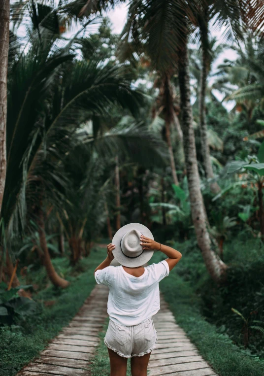 a woman walking down a path between palm trees, unsplash contest winner, sumatraism, white hat, lush garden surroundings, back view also, lush rainforest