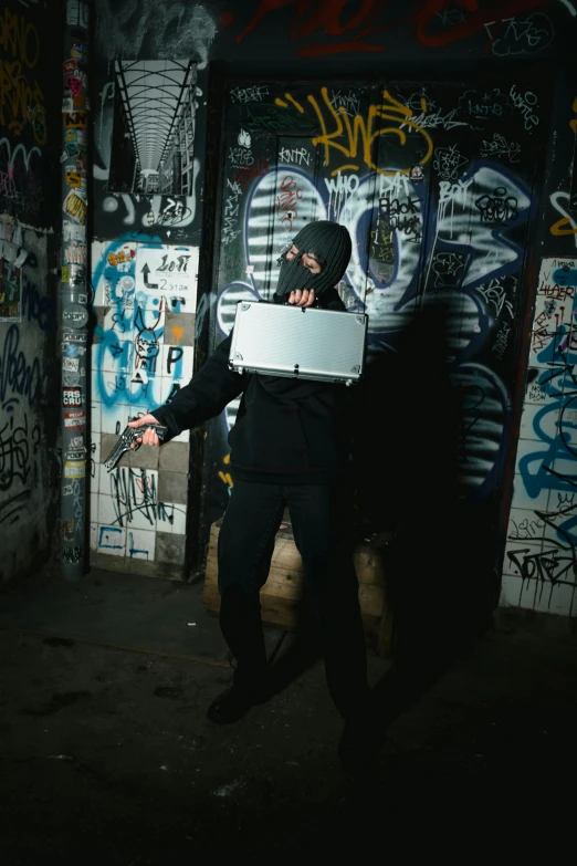 a person in a dark room with graffiti on the walls, an album cover, inspired by Banksy, unsplash, graffiti, holding glowing laptop computer, posing ready for a fight, white man with black fabric mask, holding a mp7