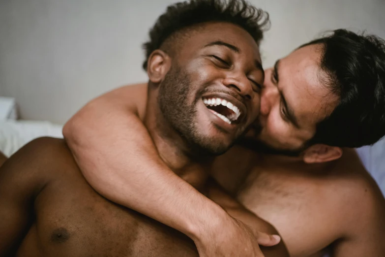 a couple of men laying on top of a bed, trending on pexels, large black smile, holding intimately, two muscular men entwined, looking across the shoulder