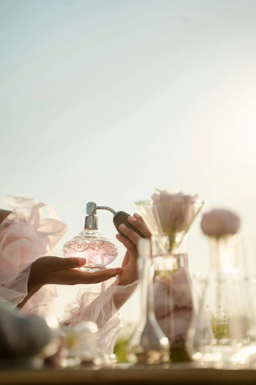 a close up of a person holding a bottle of perfume, in the sun, light pink mist, flowers, lots de details