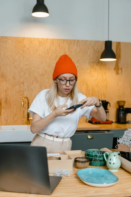 a woman sitting at a kitchen table in front of a laptop, by Julia Pishtar, beanie, scanning items with smartphone, gif, maintenance photo