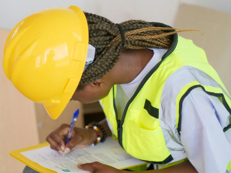 a woman in a hard hat writing on a piece of paper, by Arabella Rankin, pexels contest winner, yellow and green scheme, african sybil, still frame, well built