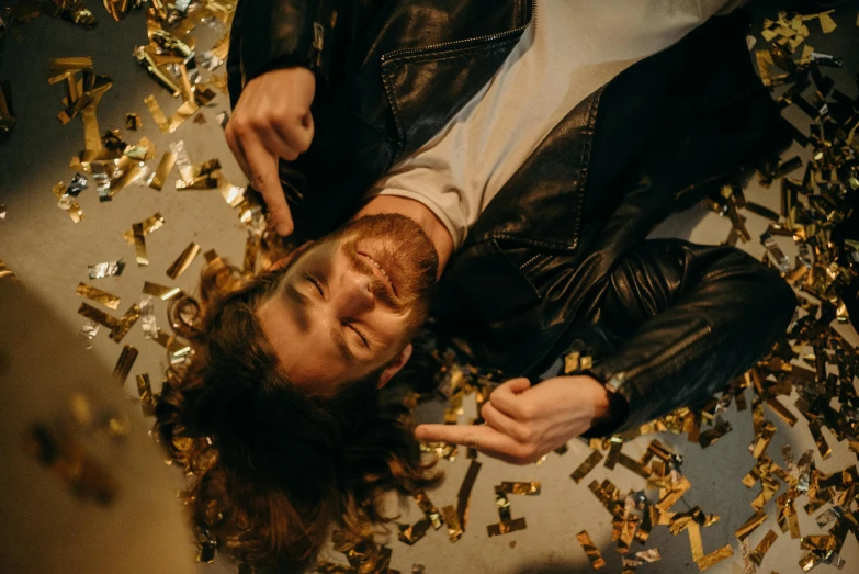 a man laying on top of a pile of gold confetti, by Julia Pishtar, trending on pexels, drunk cowboy redhead long hair, leather clothing, felix englund style, gold teeth