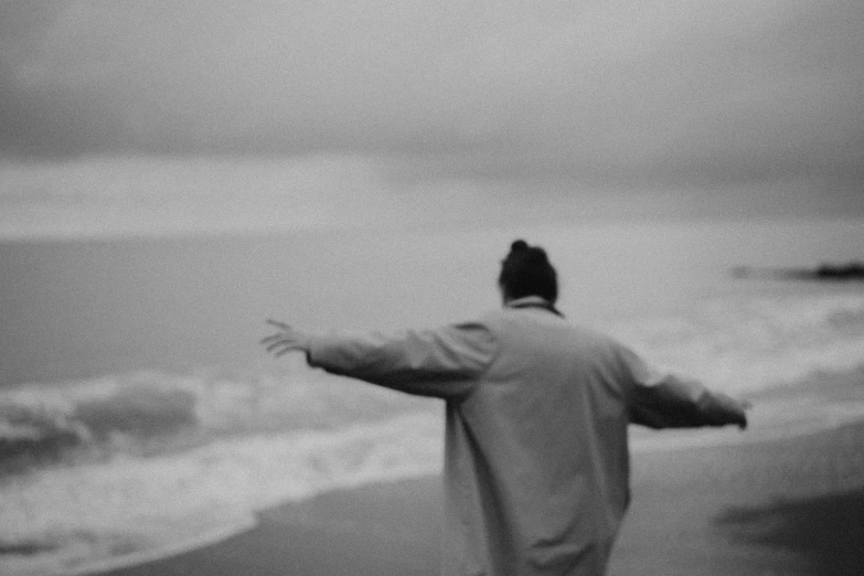 a man standing on top of a beach next to the ocean, a black and white photo, unsplash, conceptual art, woman is in a trenchcoat, waving arms, still from a music video, dream wave aesthetic