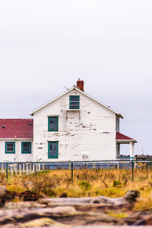 a white house sitting on top of a lush green field, a colorized photo, by Jim Nelson, unsplash, dull red flaking paint, near the seashore, white plank siding, square