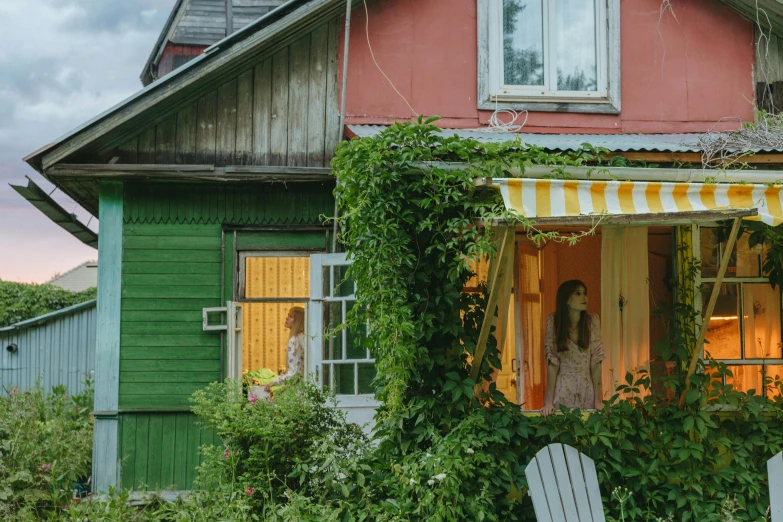 a woman standing in a window of a house, inspired by Carl Larsson, pexels contest winner, vibrant greenery outside, 3 colours, russian village, awnings