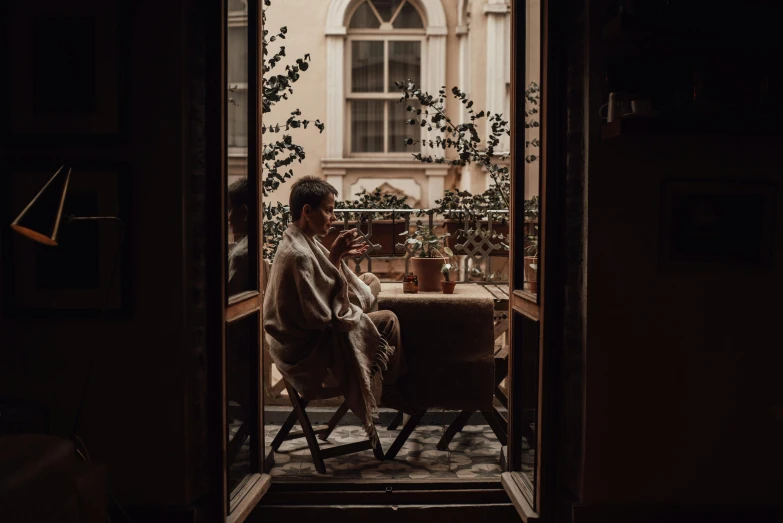 a man sitting in a chair in front of a window, by Emma Andijewska, pexels contest winner, wearing brown robes, mysterious coffee shop girl, at the terrace, spring evening