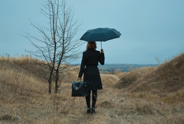 a woman walking down a dirt road holding an umbrella, an album cover, by Julia Pishtar, pexels contest winner, holding a briefcase, kramskoi 4 k, wearing black coat, blue mood